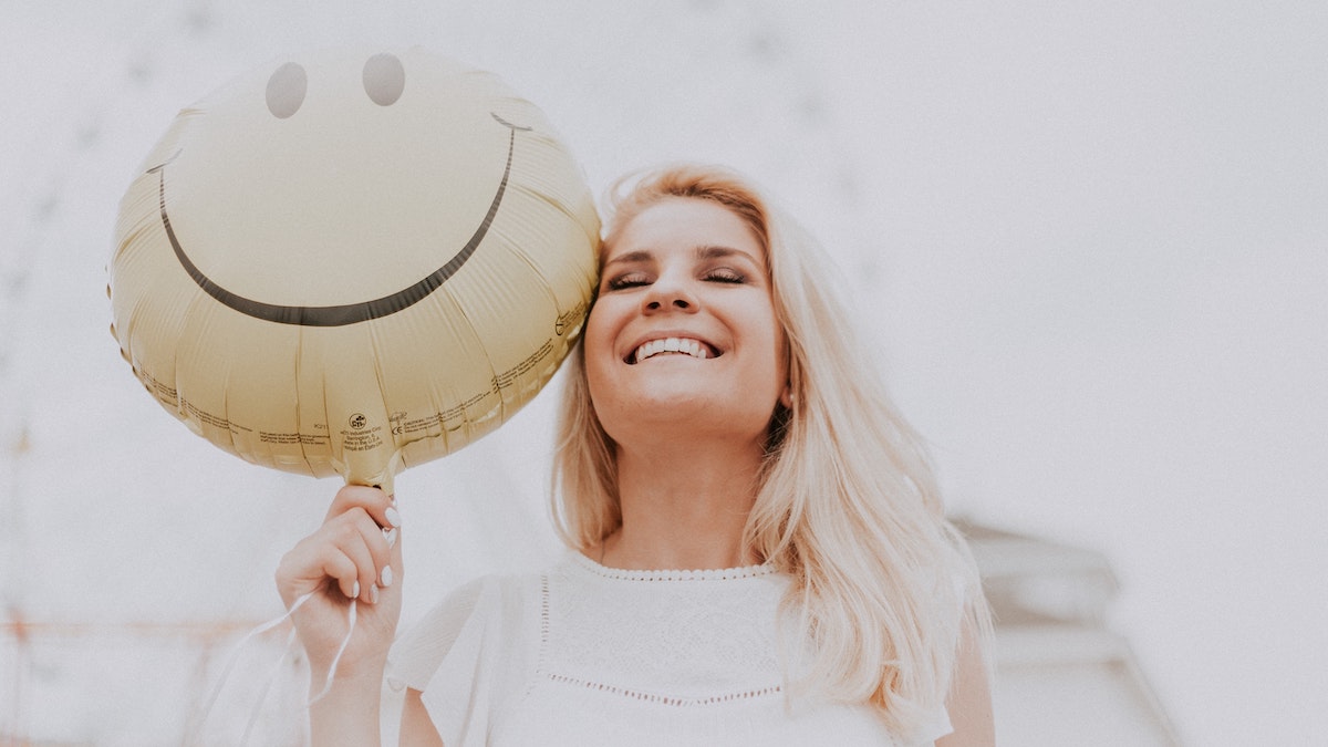 Dios sonríe: Una mujer joven rubia en una feria sonríe sosteniendo un globo amarillo de un emoticono de sonrisa.