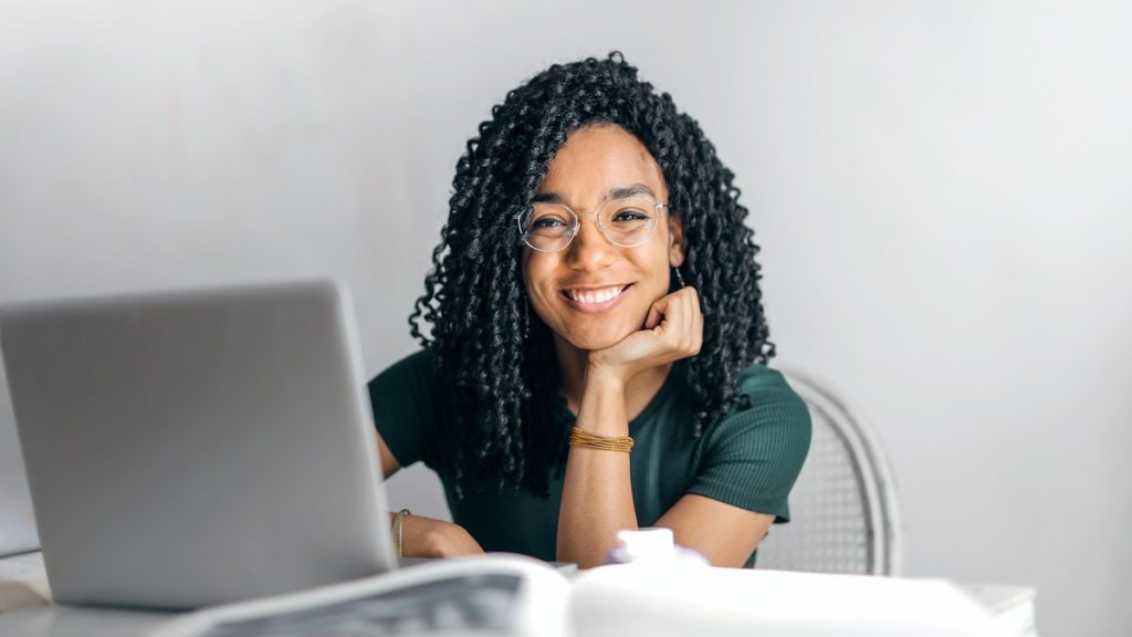 Contentamiento como estilo de vida: una joven étnica sonríe sentada delante de un portátil. Lleva gafas y una camiseta verde.