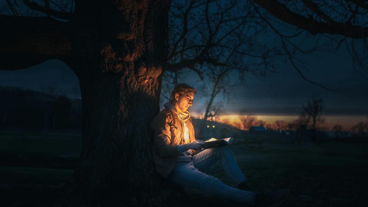 mujer iluminada en la oscuridad leyendo un libro sentada al pie de un árbol para blog sobre libros sobre Jesús en semana santa