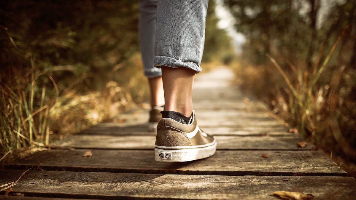 Caminar con Jesús: foto de las piernas en vaqueros o jeans desde atrás, con zapatillas de deporte marrones, caminando por una senda hecha de tablones de madera