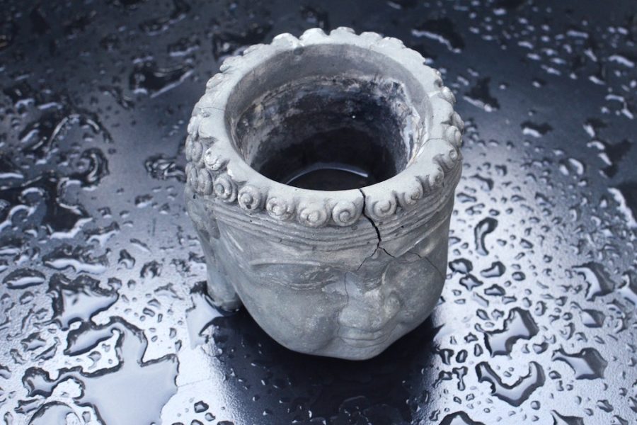 Cisternas rotas: Un vaso antiguo de piedra resquebrajado y medio vacío tallado con una cara que recuerda a un buda. Está en una superficie lisa negra llena de gotas de agua.