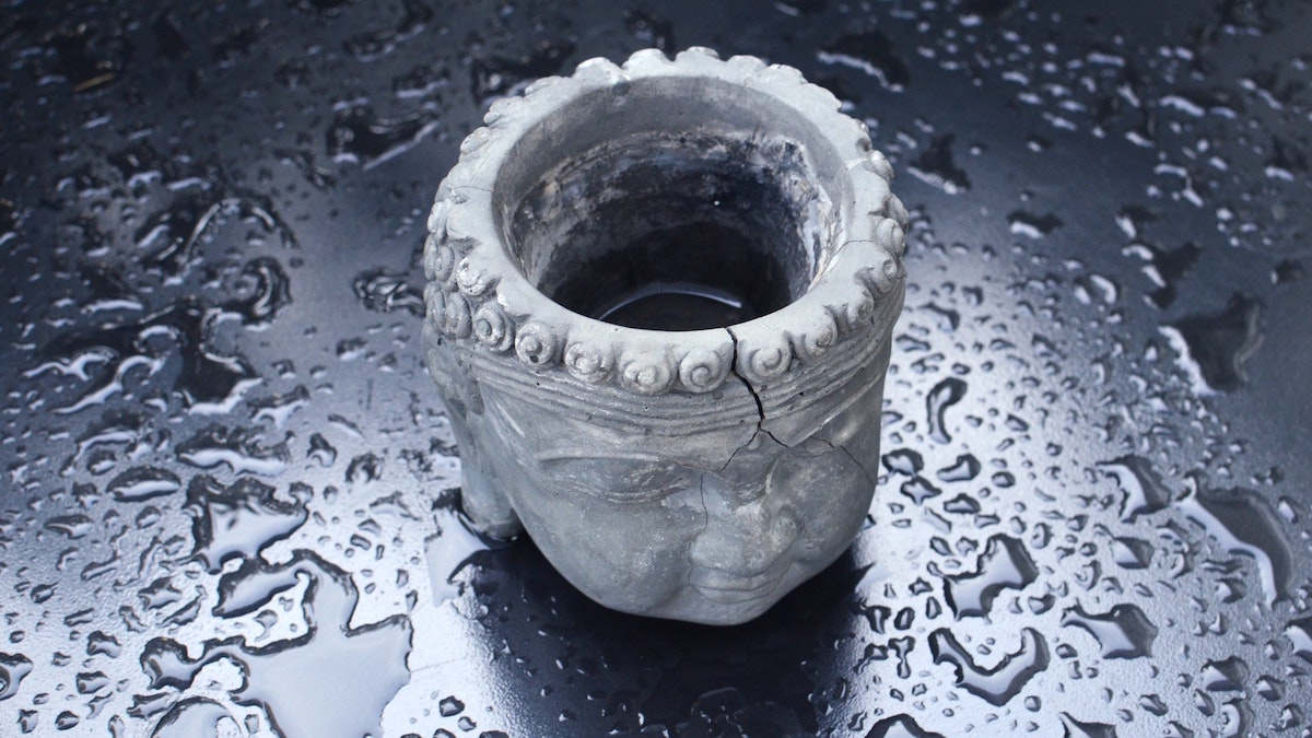 Cisternas rotas: Un vaso antiguo de piedra resquebrajado y medio vacío tallado con una cara que recuerda a un buda. Está en una superficie lisa negra llena de gotas de agua.